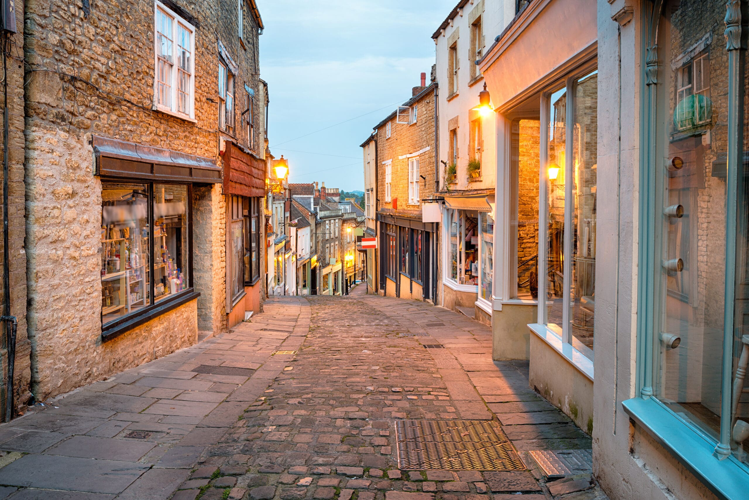 A narrow cobblestone street lined with old stone buildings and shop windows, warmly lit by street lamps at dusk—a charming setting where a skilled builder crafts tasteful house extensions in a quaint European town.